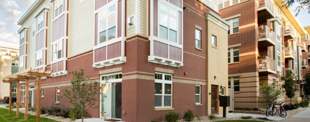 Exterior corner view of a multifamily apartment building with brick base, tan siding, and maroon accent panels, featuring a wood pergola walkway and landscaped grounds